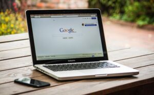 A MacBook Pro displaying Google Search on a wooden table outdoors, next to a smartphone.